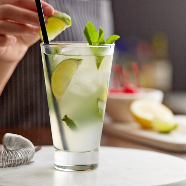 A person putting a black straw into a Libbey highball glass filled with a drink and lime slices.