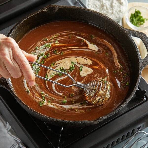 A person using a Fox Run chrome spring coil whisk to stir brown liquid in a pan.