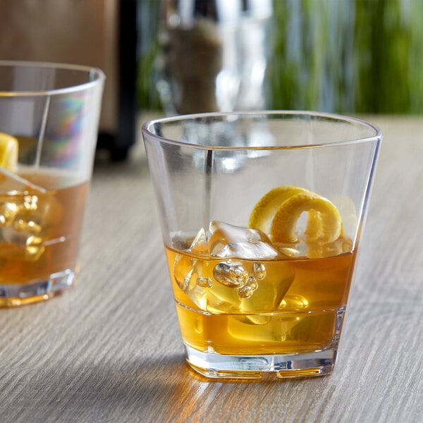 Two Libbey plastic rocks glasses with whiskey and ice on a table with a lemon peel.