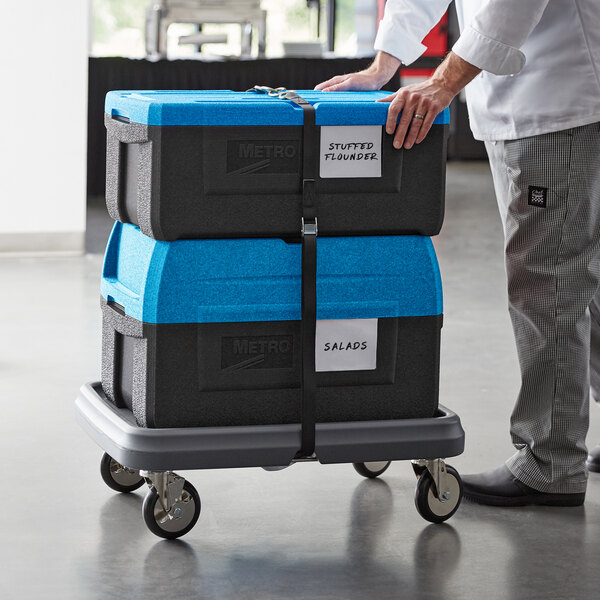 A man using a dolly to push two blue Metro Mightylite top-loading pan carriers.