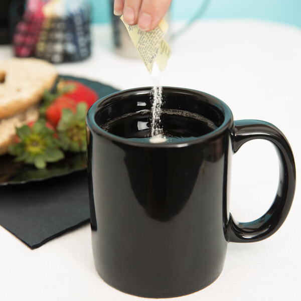 A person pouring sugar into a black Tuxton china mug.
