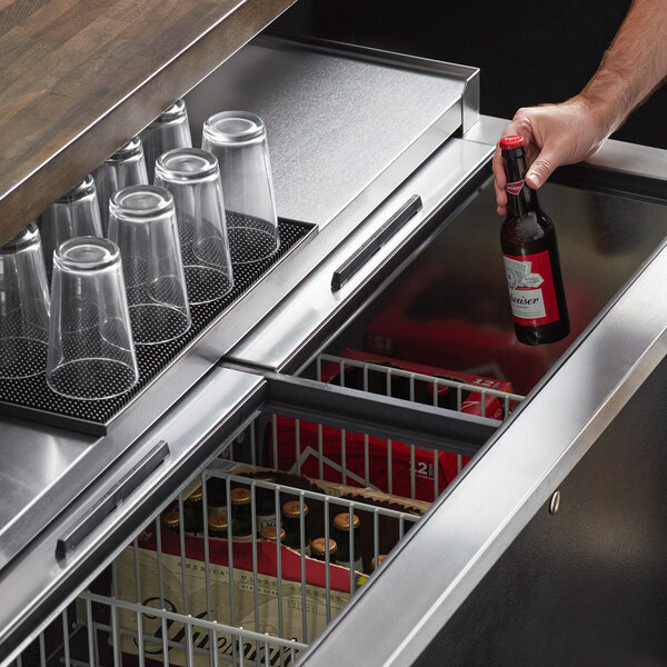 A hand holding a bottle of beer over a Beverage-Air Bottle Cooler shelf of glasses.