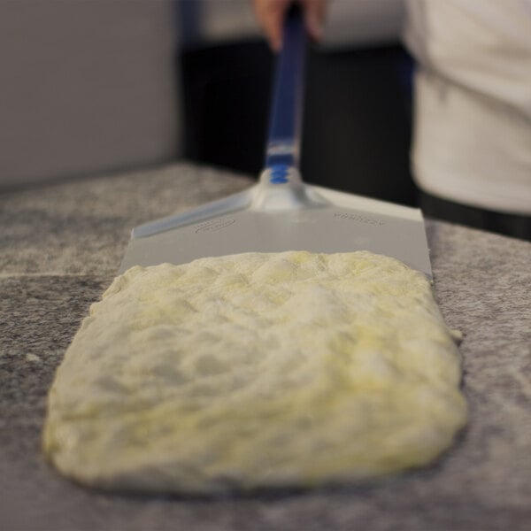 A rectangular metal pizza peel with a long blue handle is being used to lift or move a stretched pizza dough on a countertop.