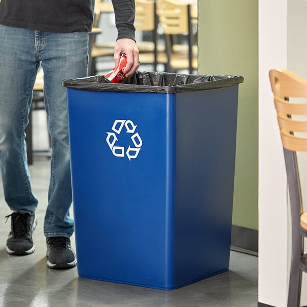 A person putting a can of soda in a blue Rubbermaid Untouchable recycling bin.