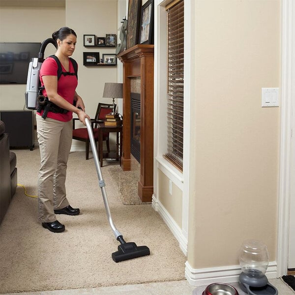 A woman using a ProTeam backpack vacuum to clean carpet in a living room.