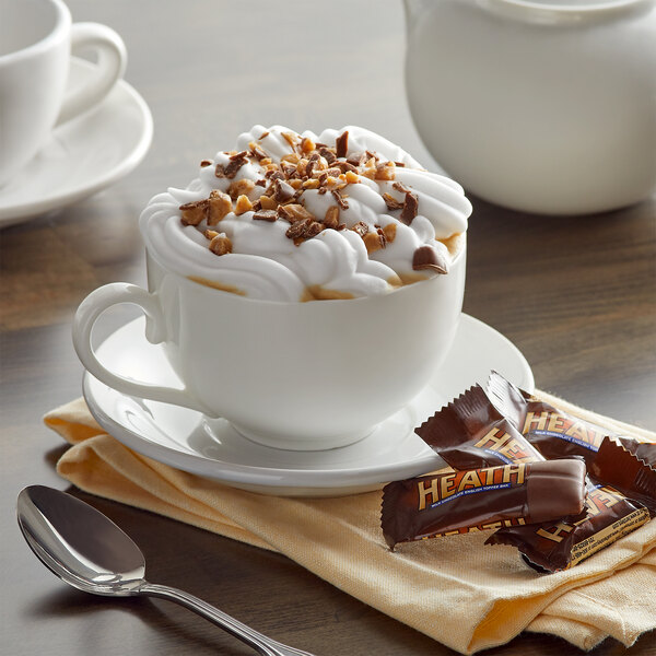 A white cup of HEATH Cappuccino with whipped cream and chocolate chips on a saucer.