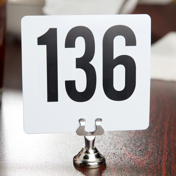 A white sign with black numbers on a stainless steel clamp-style table card holder.