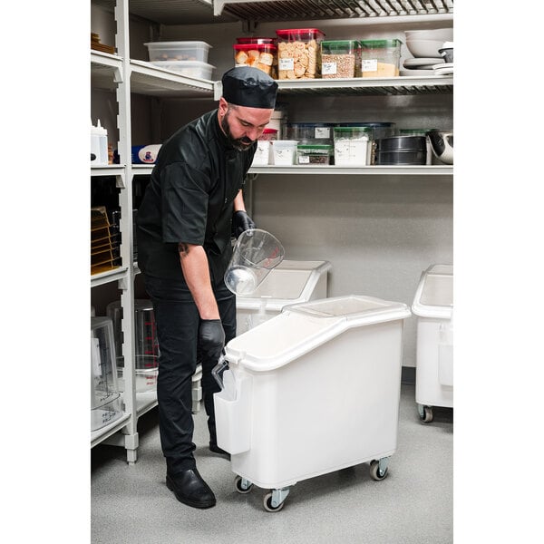 A man in a black uniform and gloves using a white Cambro ingredient storage bin on wheels.