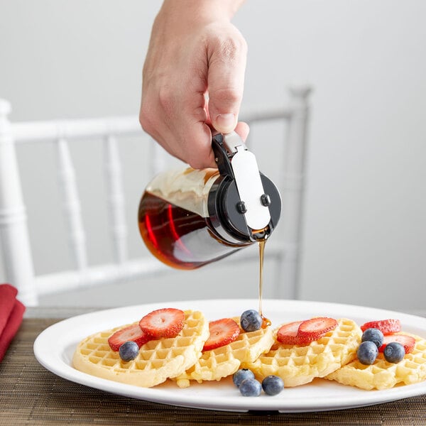 A person using a Tablecraft Modern Glass Syrup Dispenser to pour syrup on a plate of waffles with strawberries and blueberries.