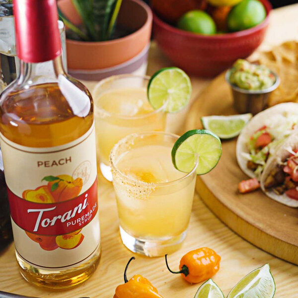 A Torani Puremade Peach Flavoring Syrup bottle on a table with a drink and lime wedges.