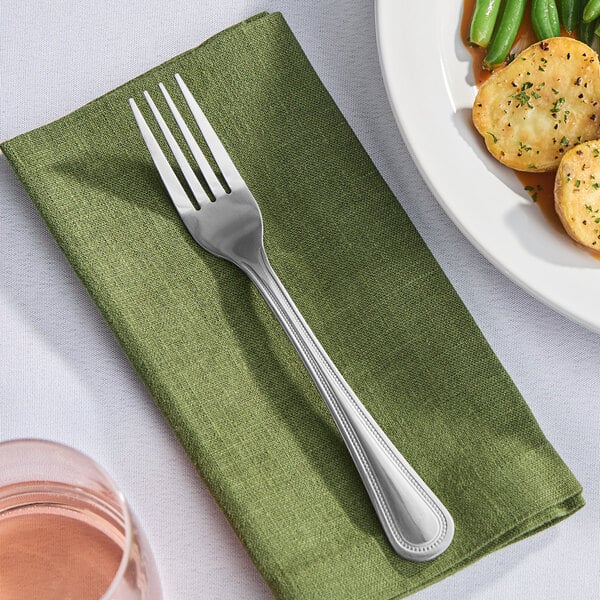 A Libbey stainless steel dinner fork on a green napkin next to a plate of food.