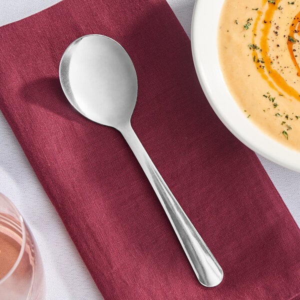 A Libbey Windsor stainless steel round bowl soup spoon on a red napkin next to a bowl of soup.