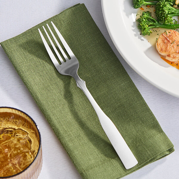 A Libbey stainless steel dinner fork on a green napkin next to a plate of broccoli.