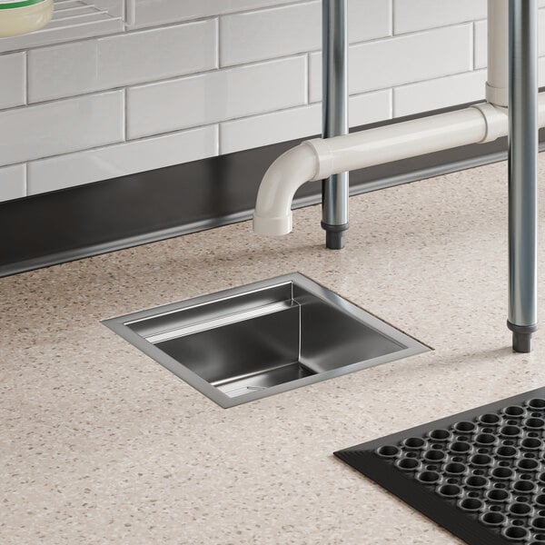 A 12-inch by 12-inch stainless steel floor sink installed in a commercial kitchen floor.