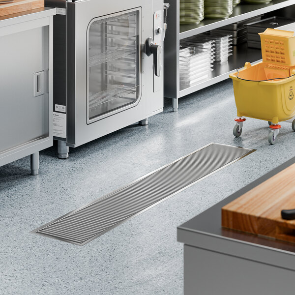 A stainless steel floor trough with a grate, measuring 12 inches by 72 inches, installed in a commercial kitchen setting.