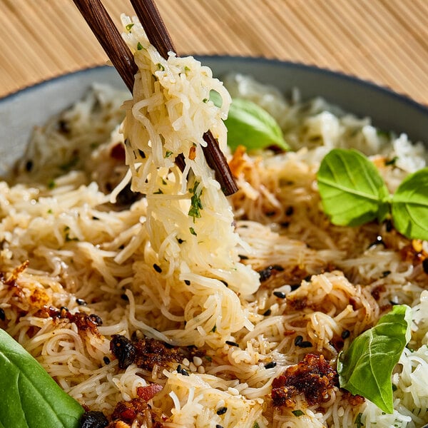 A bowl of cooked rice noodles garnished with herbs and spices, being lifted with chopsticks.