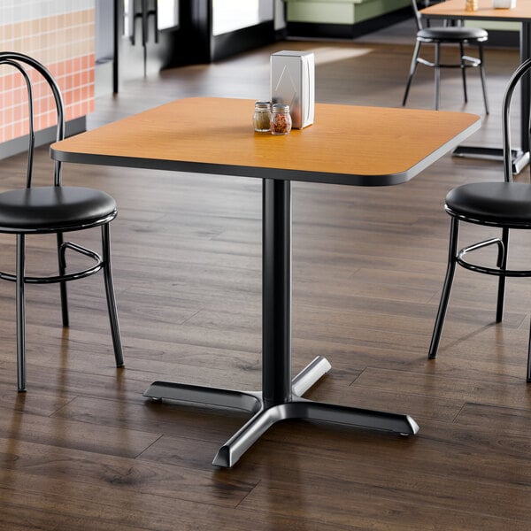 A square laminated table with a reversible walnut and oak finish, supported by a black metal pedestal base.