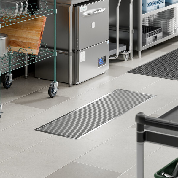 A stainless steel floor trough with a grate, measuring 18 inches by 60 inches, installed in a commercial kitchen setting.