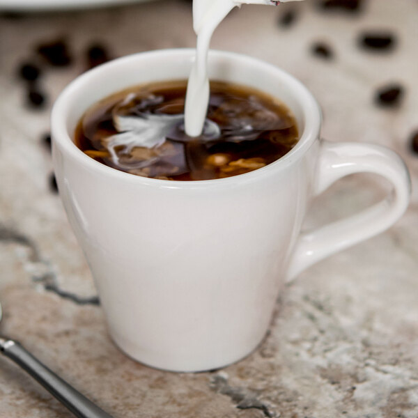 A white Tuxton espresso cup filled with white liquid and more being poured in.