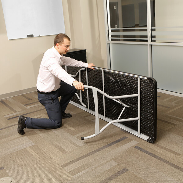 A man kneeling on his knees setting up a Lifetime black plastic folding table in a room.