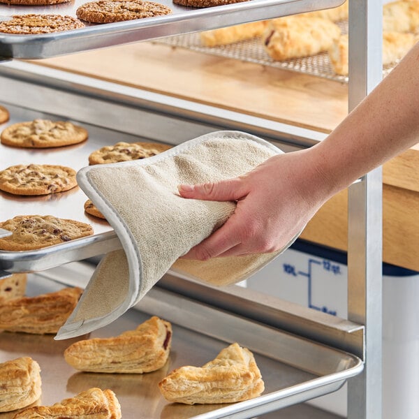 A beige terry cloth pan grabber or baker's pad being used to handle a metal baking tray with cookies and pastries.
