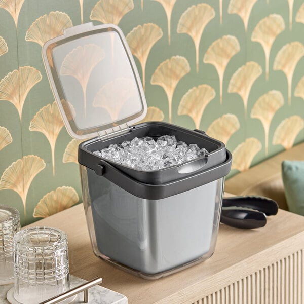 An OXO Good Grips plastic ice bucket with tongs and garnish tray filled with ice on a table.