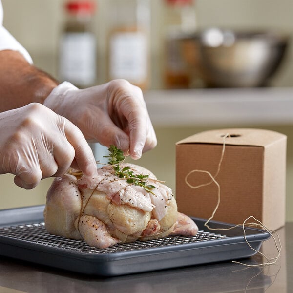 Gloved hands tying an herbed chicken on tray with boxed linen twine