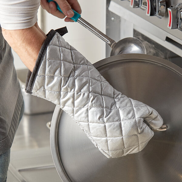 A person wearing silver silicone-coated oven mitts holding a ladle over a pot.