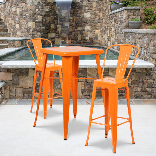 An orange metal bar height table with two chairs on an outdoor patio by a fountain.