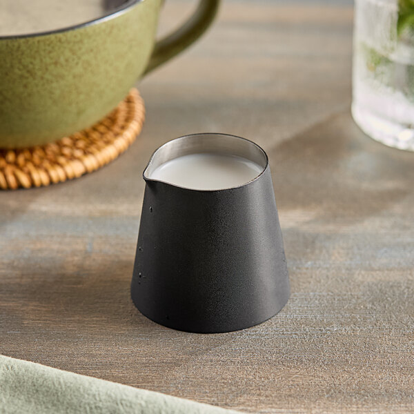 An American Metalcraft black satin stainless steel creamer with white liquid on a table.