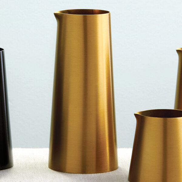 An American Metalcraft gold satin stainless steel creamer on a table with black vases.