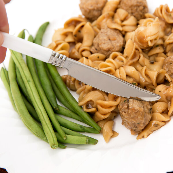 A WNA Comet Reflections Duet stainless steel look plastic knife in white on a plate of pasta with meatballs and green beans.