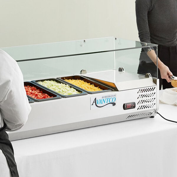 A woman in a white uniform standing behind an Avantco glass sneeze guard serving food on a counter.