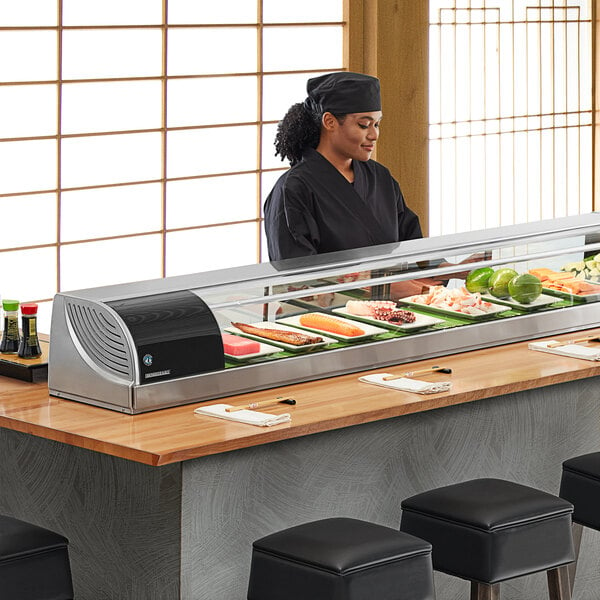 A woman standing behind a Hoshizaki refrigerated sushi display case full of sushi.