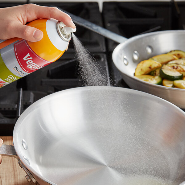 A person spraying a silver pan of vegetables with Vegalene Olive Mist using a spray can.
