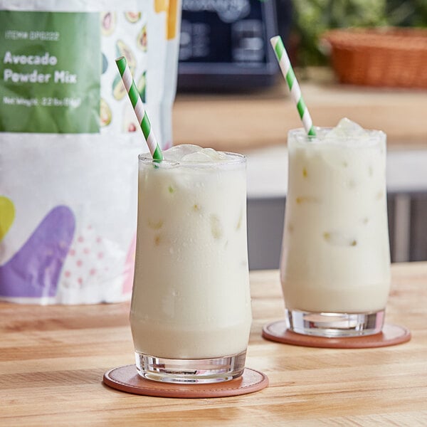 Two glasses of avocado milkshakes with green and white striped straws on a table.