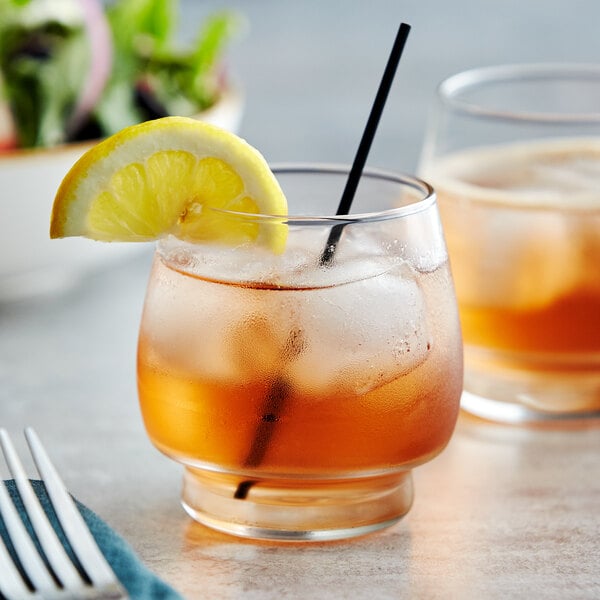 A Libbey stackable rocks glass with ice and a lemon slice on a table with a blue cloth and a glass of liquid.