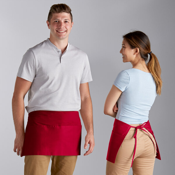 A man and woman wearing Choice red customizable waist aprons over white shirts standing in a professional kitchen.