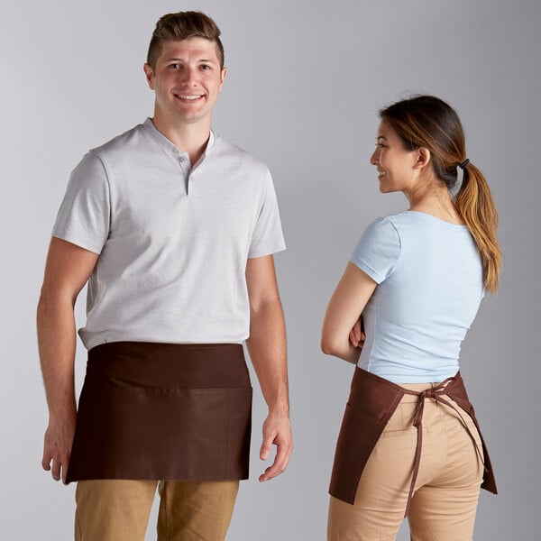 A man and woman standing next to each other wearing brown Choice standard waist aprons with 3 pockets.