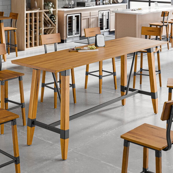 A rectangular solid wood bar height table with a honey maple finish and trestle base, featuring industrial metal accents.