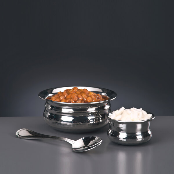 A silver World Tableware stainless steel pot belly bowl filled with beans and onions with a spoon on the table.