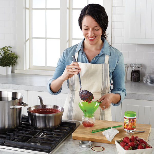 A woman in an apron mixes strawberries in a green bowl next to a Ball glass jar filled with red liquid.