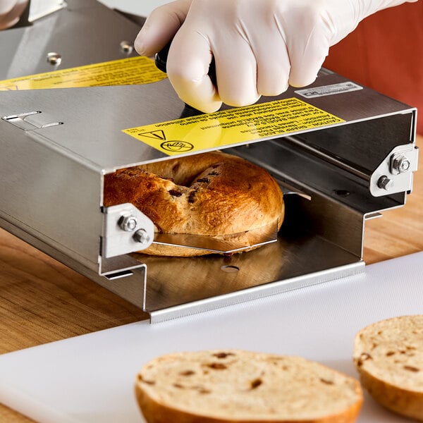 A person wearing gloves is using a metal bagel slicer to cut a bagel in half on a wooden surface.