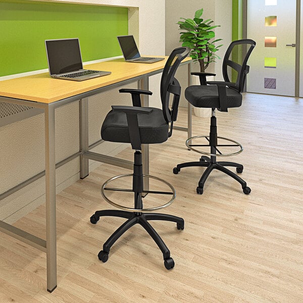 A black Boss drafting stool with a metal base and footring at a desk with a laptop.