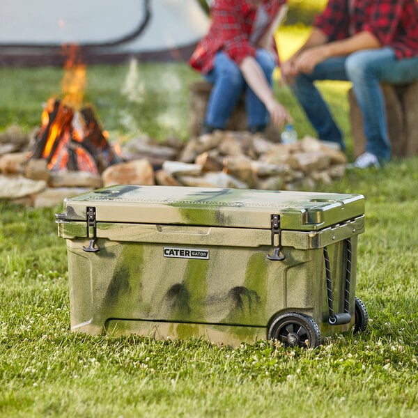 A CaterGator camouflage cooler on grass with people sitting around it.