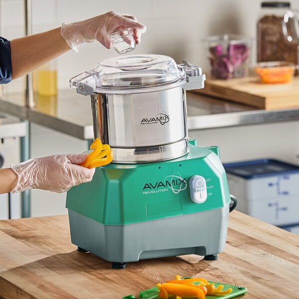 A stainless steel batch bowl food processor with a green and gray base labeled 'AvaMix Revolution' being used to process sliced yellow peppers.