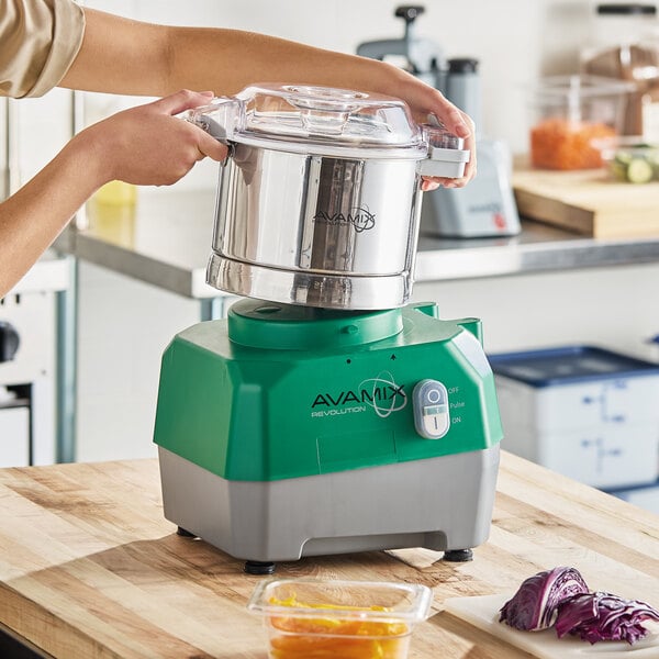A green and gray AvaMix Revolution combination food processor with a stainless steel bowl being placed on its base in a kitchen setting.