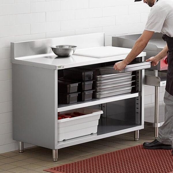 A man in a white shirt and black pants standing next to a Regency stainless steel enclosed base work table with a backsplash.