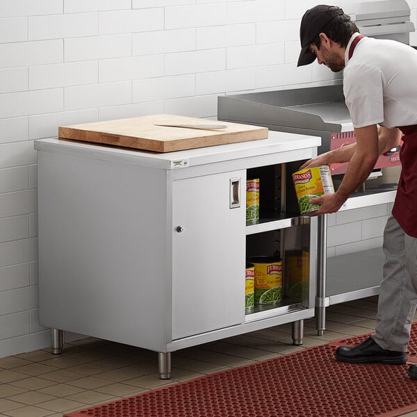 A man in a black apron standing in front of a Regency stainless steel enclosed base table with sliding doors and adjustable midshelf.