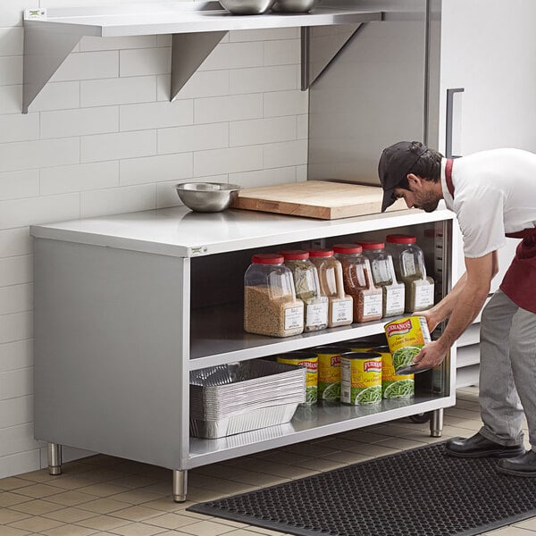 A man in a professional kitchen using a stainless steel table with a shelf full of food.